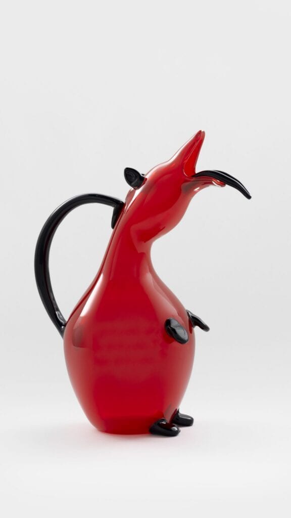 A red and black glass pitcher shaped like an abstract animal with a long snout, upright posture, and small limbs, set against a plain white background.