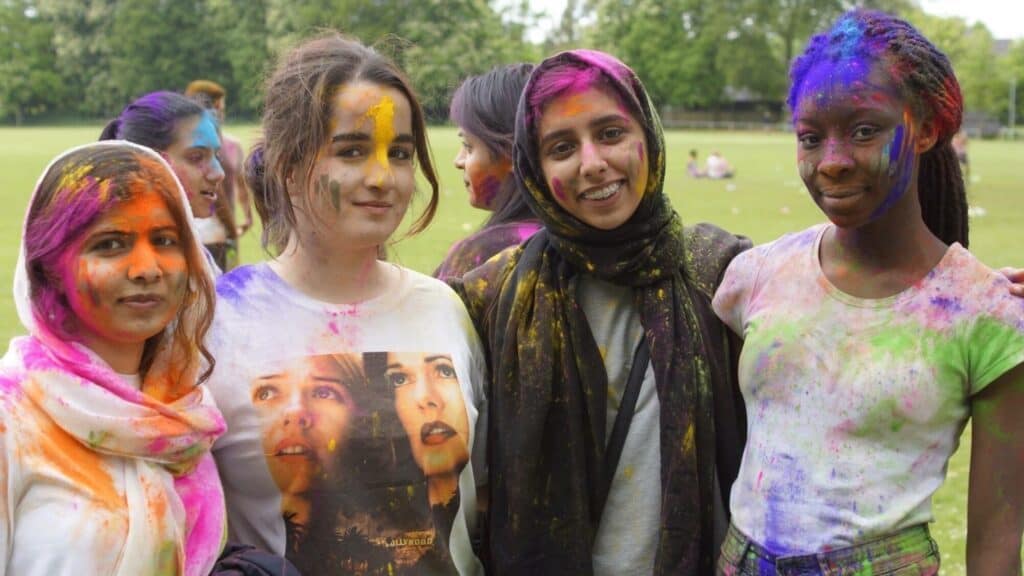 Four young women stand outdoors with colorful powder on their faces and clothes, smiling at the camera during a festival, with others and greenery visible in the background.