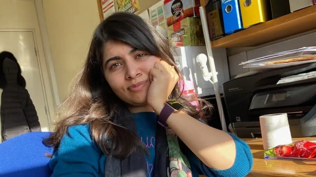 A woman with long dark hair sits at a desk, resting her face on her hand. Office supplies, folders, and a printer are visible in the background. Sunlight shines through a window as she reads Malala Yousafzai news on her computer.