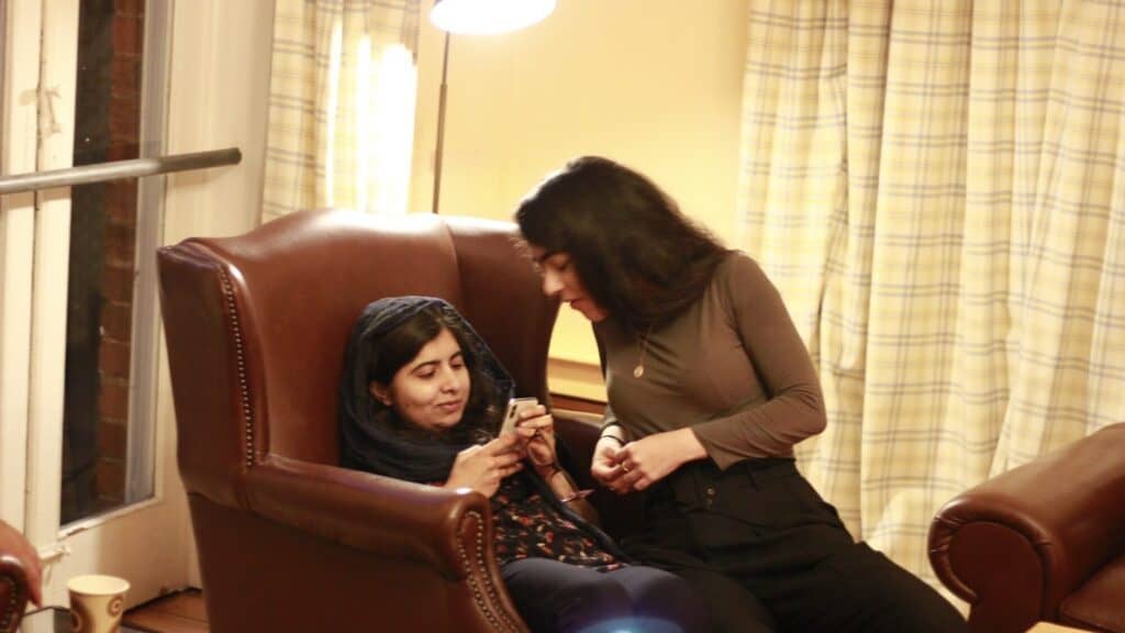 Two women sit together in a cozy room; one reclines in a leather chair looking at her phone while the other leans in, engaging with her. A lamp and curtains are in the background.