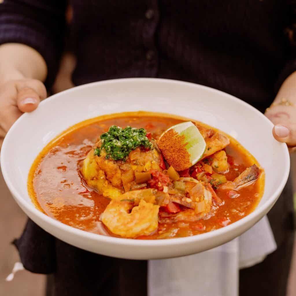 A person holds a white bowl filled with seafood stew, including shrimp, fish, vegetables, a lime wedge, and green garnish in a red broth.