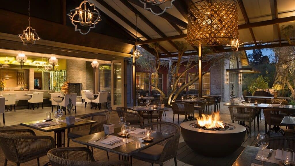 Upscale restaurant patio with wicker chairs, covered roof, tables set for dining, and a large fire pit in the center; indoor dining area visible through glass doors.