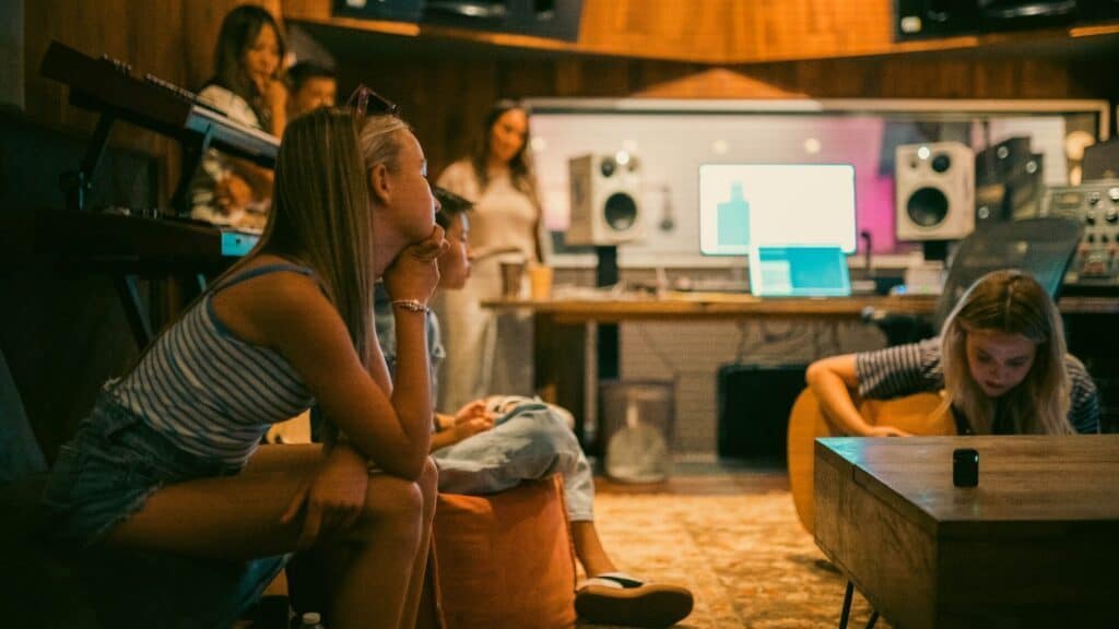 Several people in a recording studio; one person plays guitar, another sits listening, while others stand and chat near sound equipment and a large monitor.