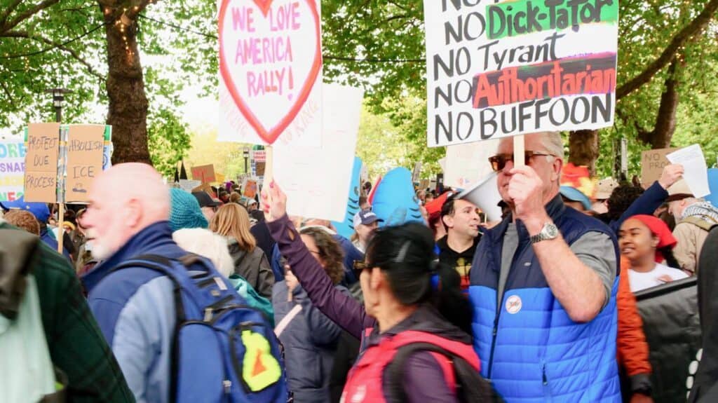 A crowd of protesters gathers outdoors holding various signs, including ones reading "WE LOVE AMERICA RALLY" and "NO Dictator, NO Tyrant, NO Authoritarian, NO BUFFOON.