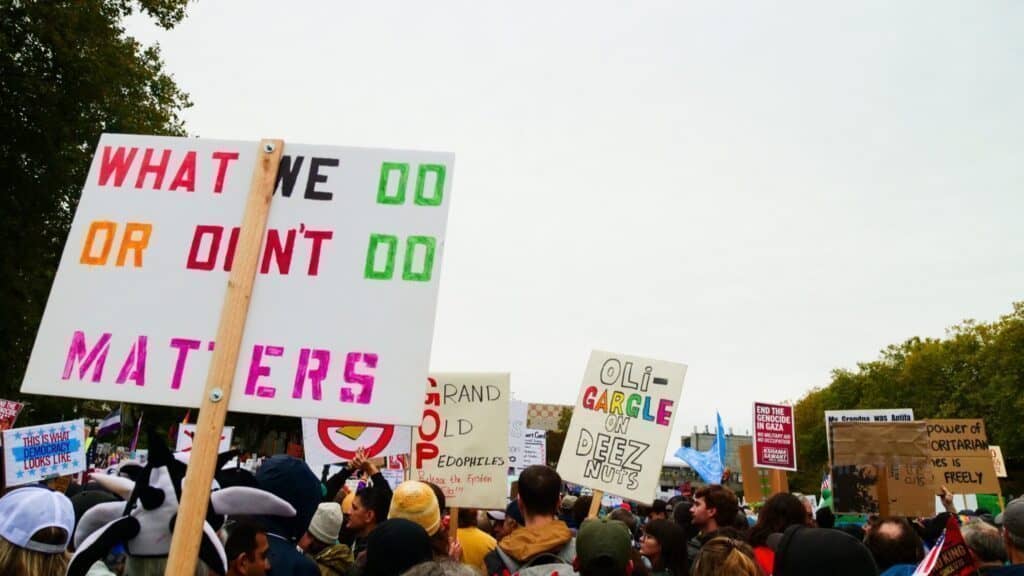A crowd of people at a protest hold up various handmade signs with slogans, including one that reads “WHAT WE DO OR DIDN’T DO MATTERS.” Trees and cloudy sky are visible in the background.