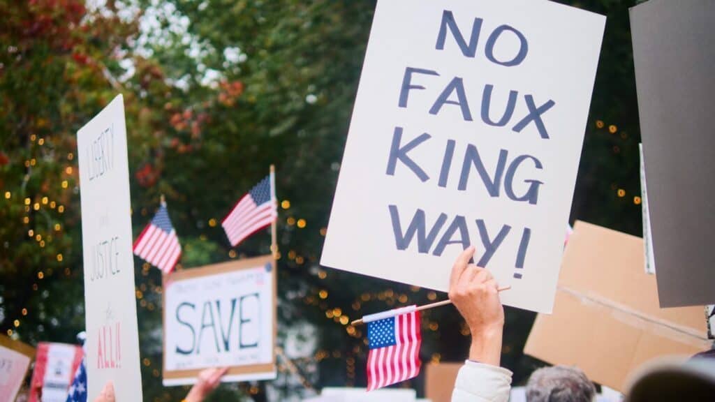 A protester holds a sign that reads "NO FAUX KING WAY!" among a crowd with American flags and other signs. Trees and lights are visible in the background.