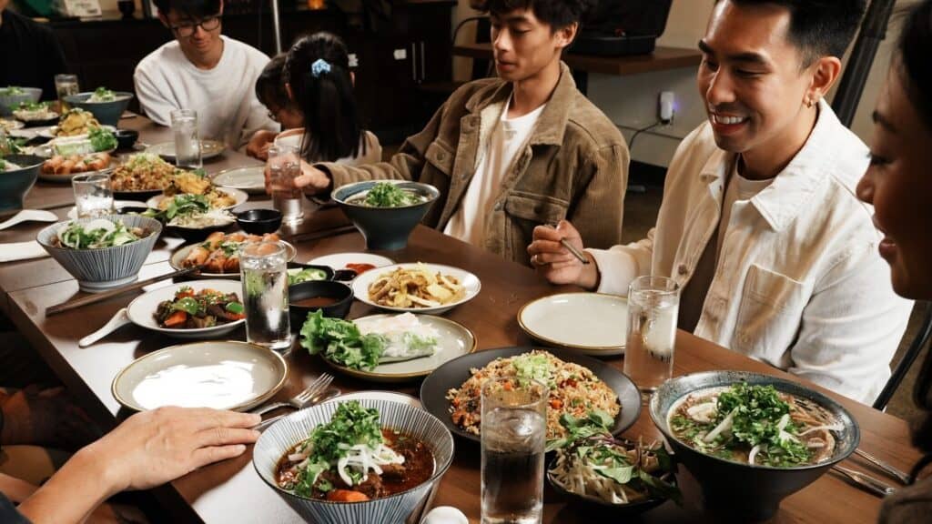 A group of people sit around a dining table filled with various Asian dishes, including noodles, salads, and greens, sharing a meal together.