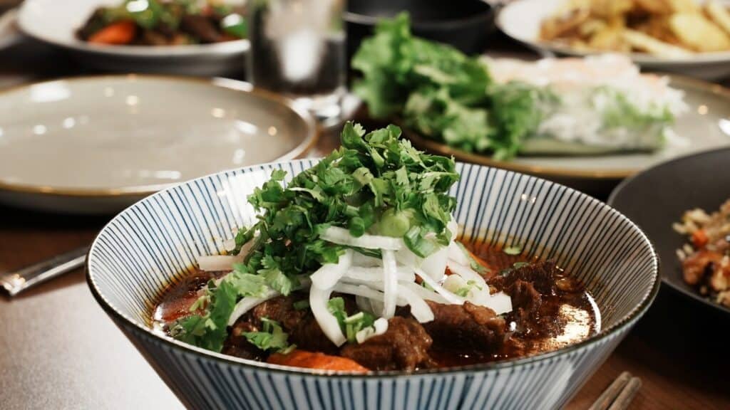 A bowl of beef stew garnished with chopped cilantro and sliced onions, with various plates of food and greens in the background on a dining table.