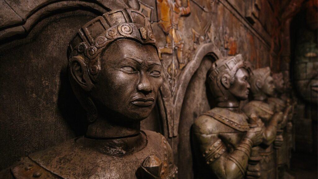A row of ancient stone statues with detailed headpieces and armor, set against a textured wall with carvings.