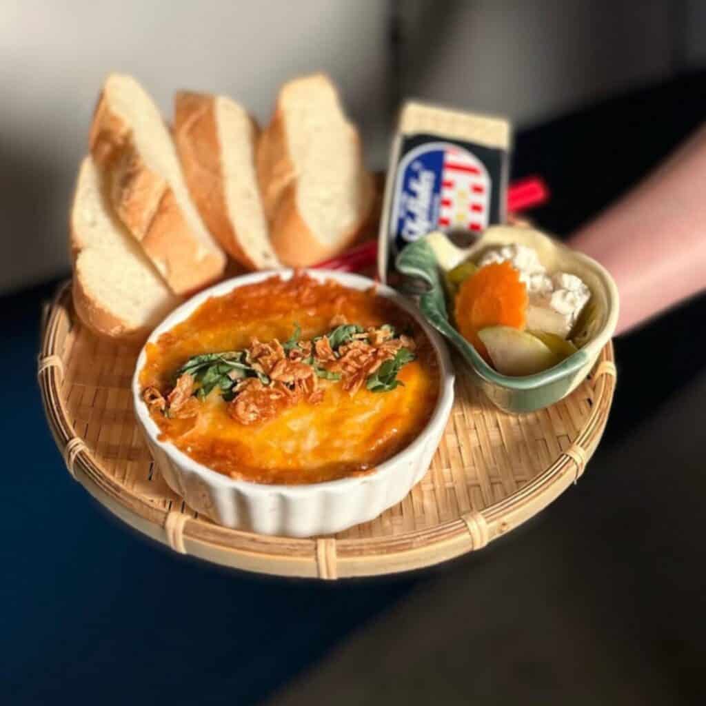 A round tray with a baked dish topped with herbs, four slices of bread, a small bowl of pickled vegetables, and a pack of processed cheese.