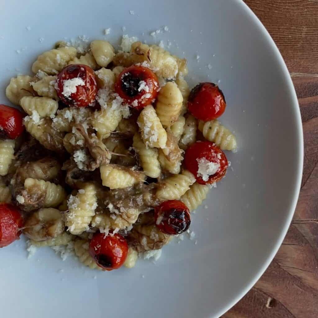 A plate of gnocchi topped with shredded meat, roasted cherry tomatoes, and grated cheese on a white dish—perfect October food for those seeking comforting things to eat.