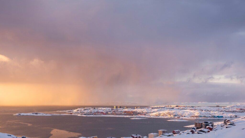 A coastal town with snow-covered buildings and landscape under a dramatic, cloudy sky with warm sunlight on the horizon.
