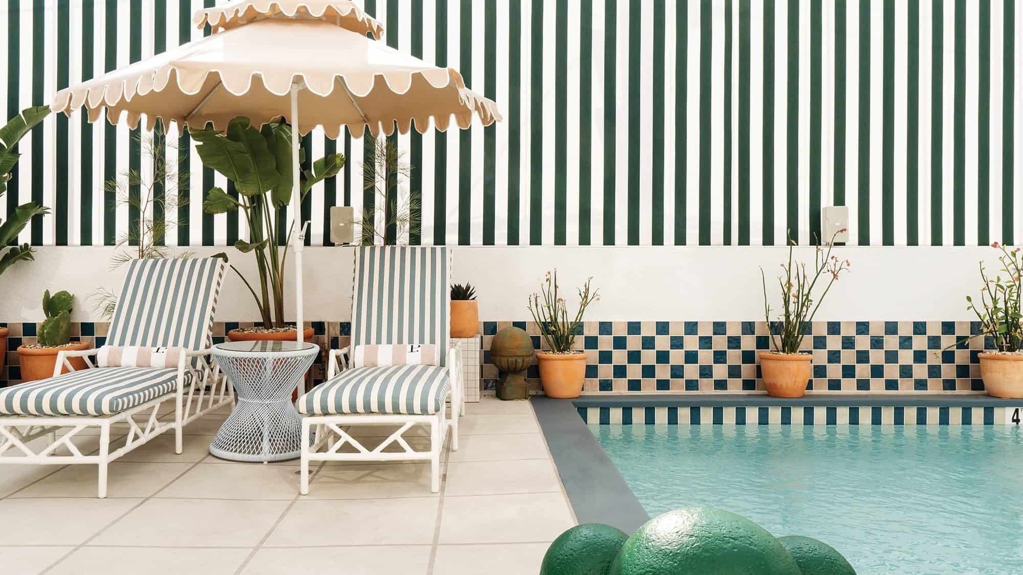 Two striped lounge chairs and a scalloped umbrella sit by a pool in Los Angeles, with potted plants and a green-and-white striped wall in the background.