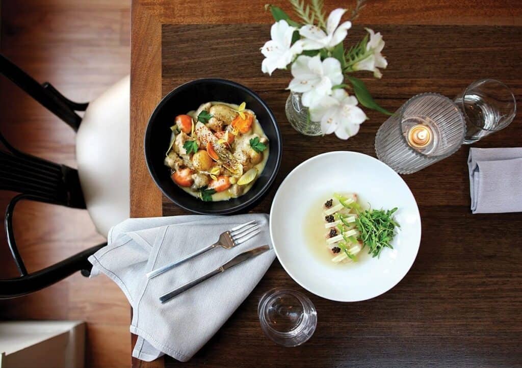 Overhead view of a table set with two plated dishes, a napkin with cutlery, a glass of water, a candle, and a small vase of white flowers—capturing the dining charm you’ll find while visiting LA, the city of Angels.