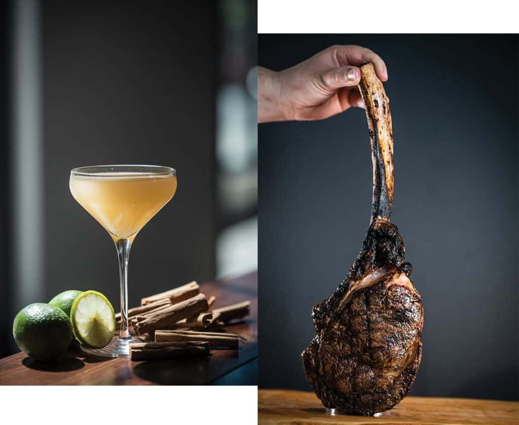 A cocktail in a coupe glass with limes and cinnamon sticks beside it, next to a hand holding a large, grilled tomahawk steak—a taste of small towns and big flavors you might find on your travels through Montana.