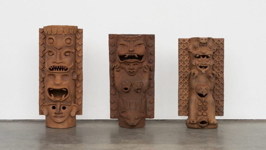 Three brown, sculpted totem-like figures with intricate, abstract facial and body features stand upright against a plain white wall on a concrete floor.