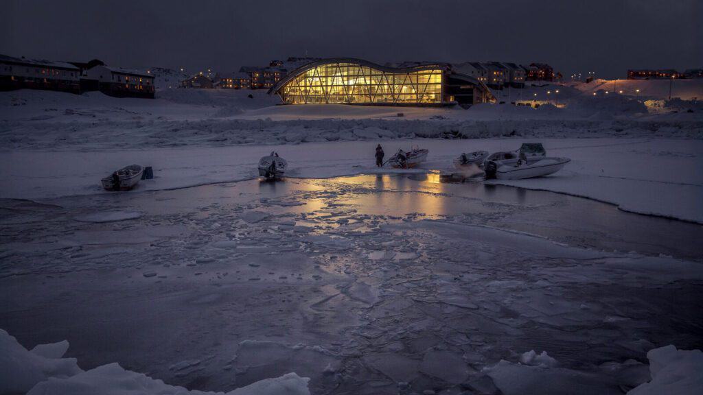 Boats are docked in a partially frozen body of water at night, with a modern, illuminated building and snow-covered landscape in the background.