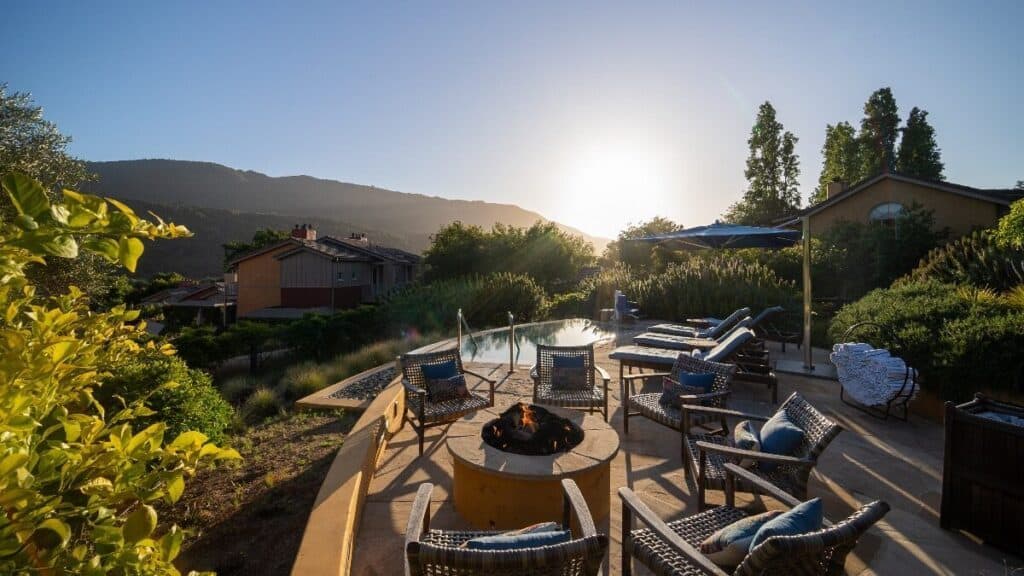Outdoor patio with wicker chairs around a fire pit, lounge chairs by a pool, and houses with mountains in the background at sunset.