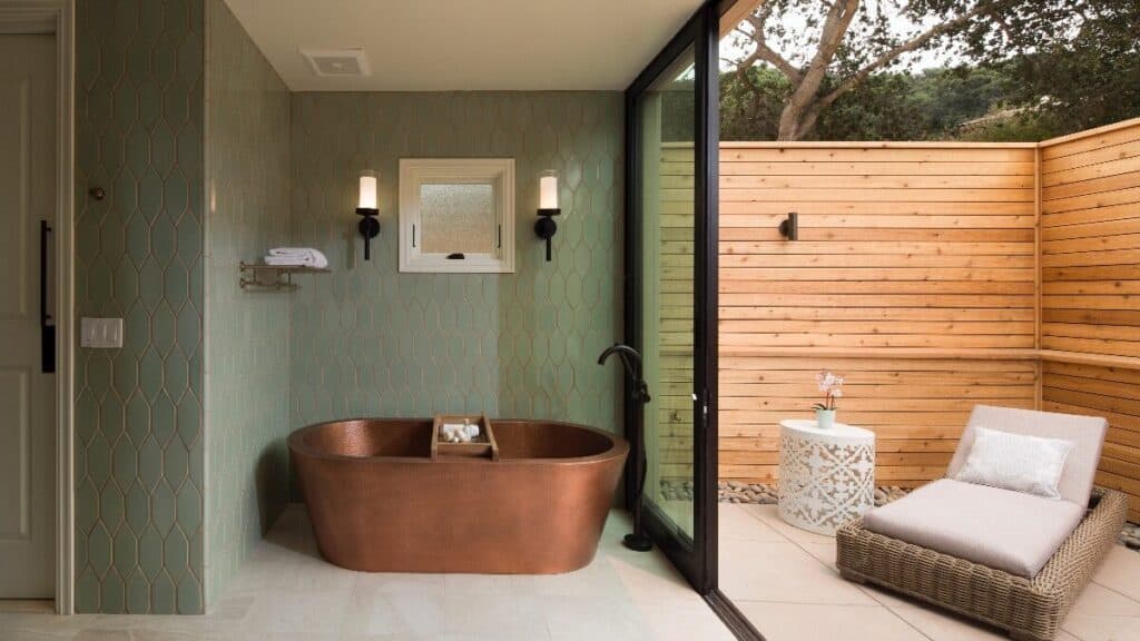 A modern bathroom with a copper bathtub, wall-mounted lights, and sliding glass doors opening to an outdoor patio with a lounge chair and small table.