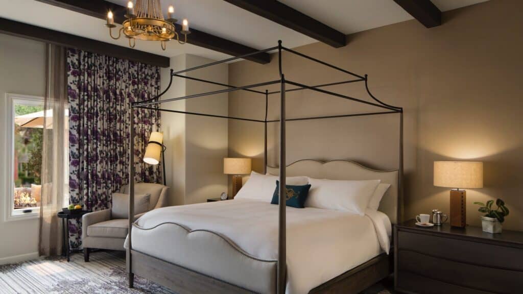 A well-lit bedroom with a canopy bed, two bedside tables with lamps, an armchair by the window, and floral curtains.