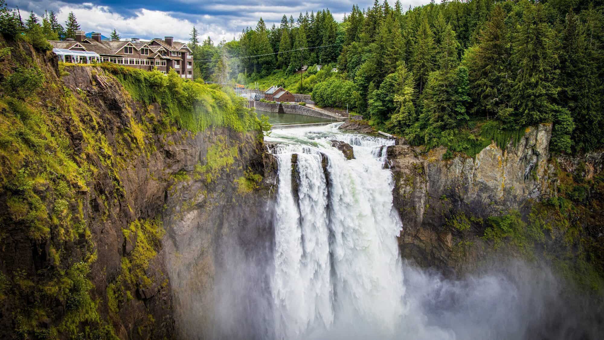 A large waterfall cascades over a rocky cliff surrounded by dense green forest in Washington, with a lodge building at the cliff's edge, reflecting the area's Native Heritage.