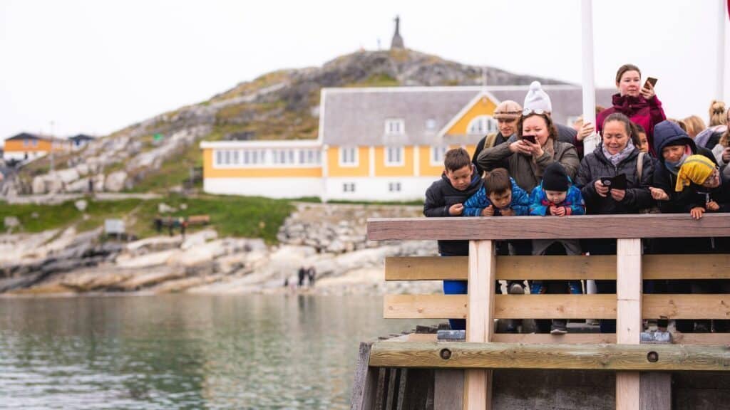 A group of people, including several children, stand on a wooden dock in Nuuk, Greenland, looking down at the water, with buildings and a hill in the background.