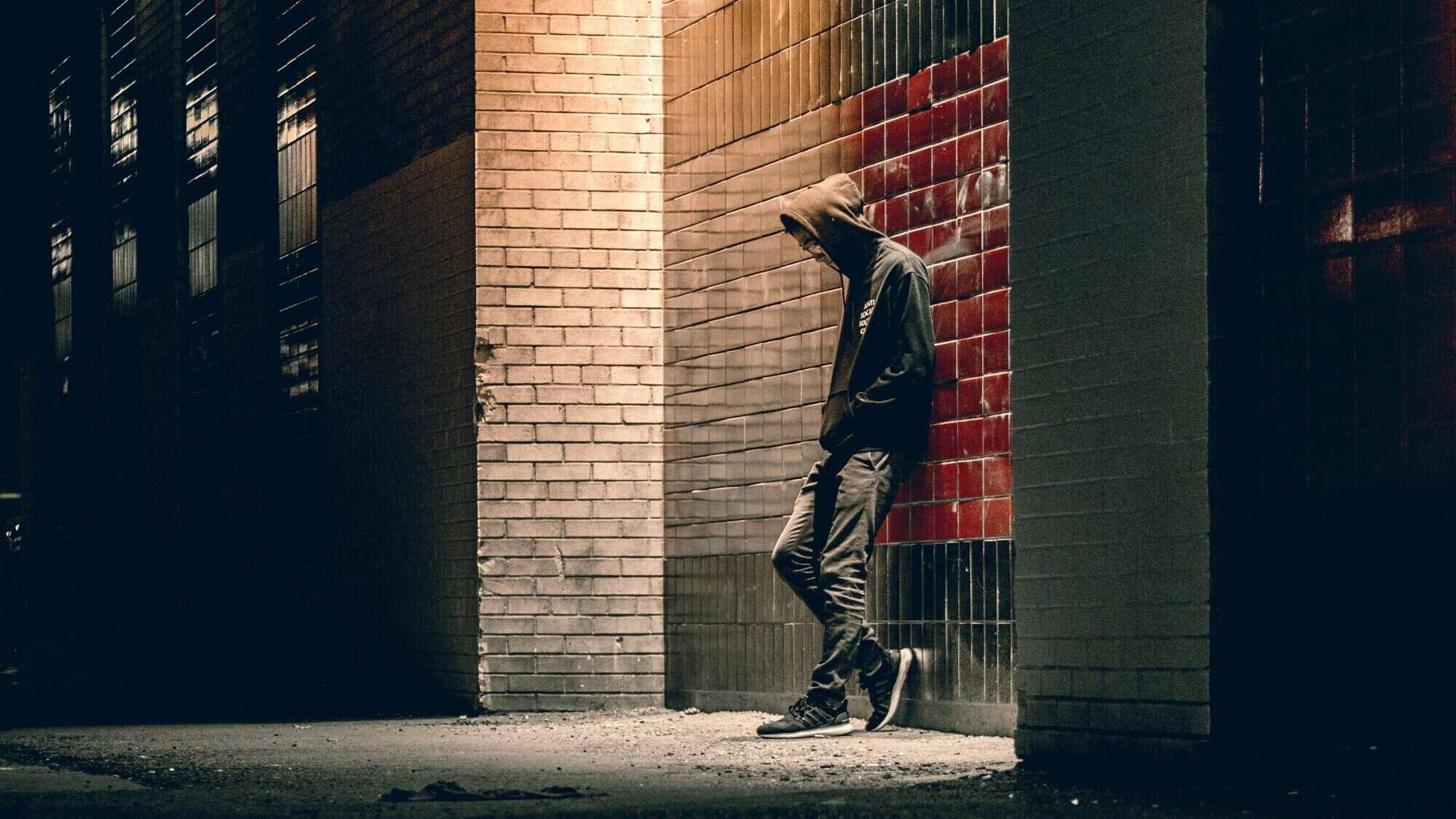 A person in a hoodie stands alone at night, leaning against a brick wall with red tiles, partially illuminated by a streetlight, capturing the quiet grief and boys' pain in the stillness.