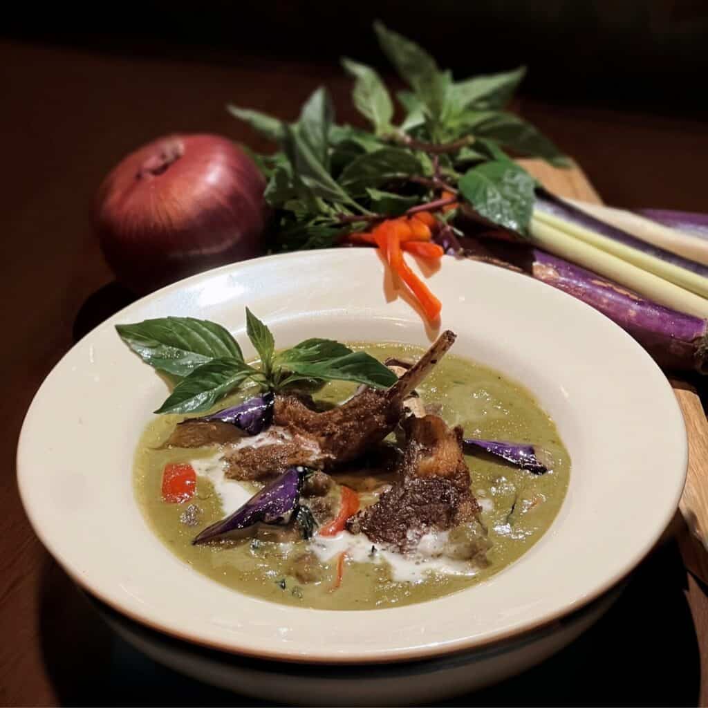 A bowl of green curry with lamb chops, garnished with basil, sliced eggplant, and red chilies—one of November’s must-try dishes—set against a background of raw onion, basil, chili, and lemongrass.