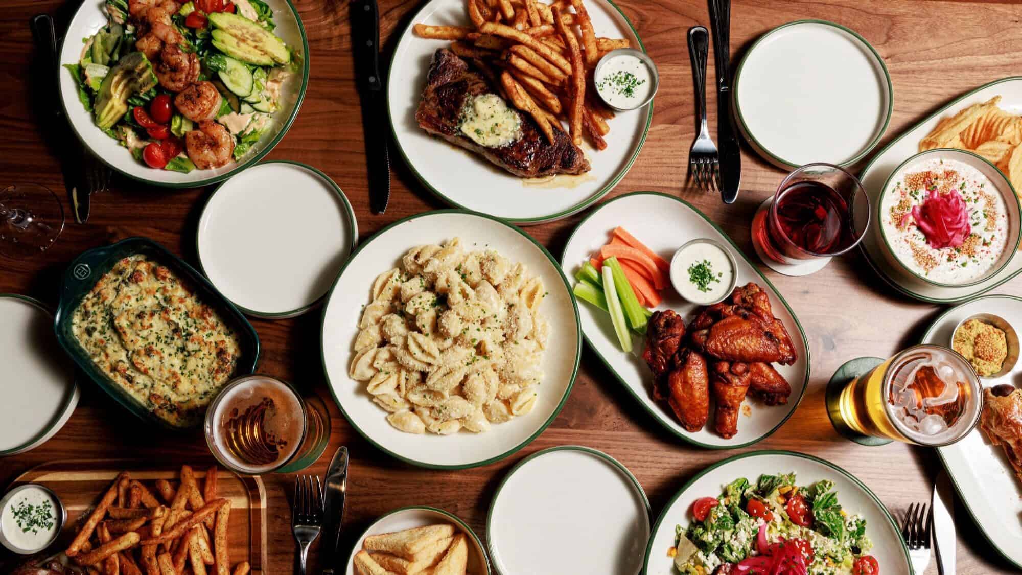 A table set with plates of steak and fries, pasta, chicken wings, salads, dips, drinks, and side dishes on a wooden surface.