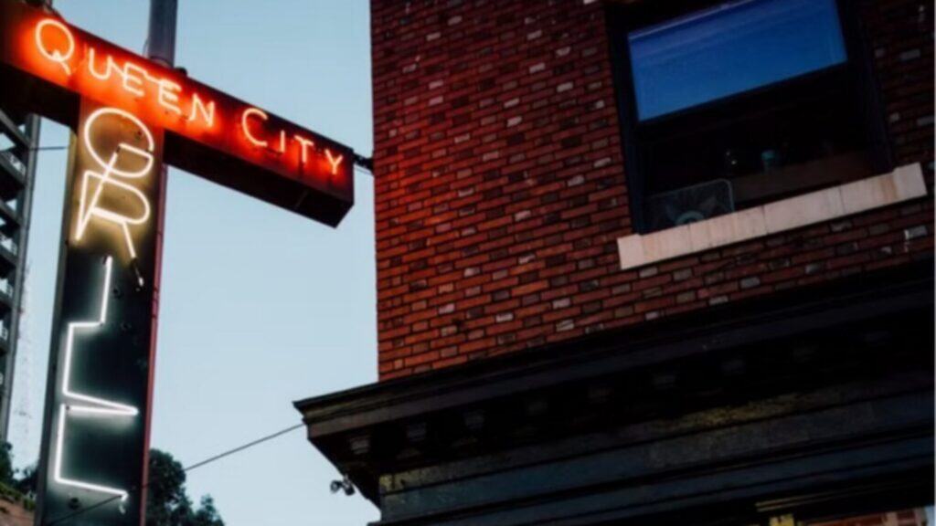 A neon sign reading "Queen City Grill" glows on the side of a red brick building in Belltown during dusk, highlighting the legacy of this Queen City landmark.