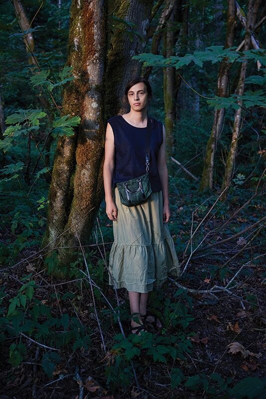 A person wearing a sleeveless black top and a light green skirt stands against a tree in a forest, looking at the camera—captured by Seattle photographer Camille Trautman.