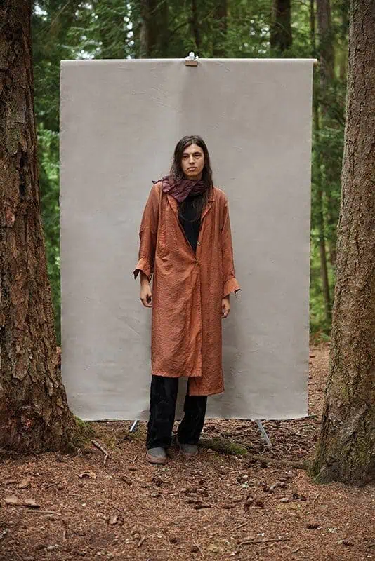 A person in a long rust-colored coat stands in front of a plain backdrop between two trees in a forest, evoking the introspective style of Camille Trautman from the Boren Banner Series at the Frye Art Museum.