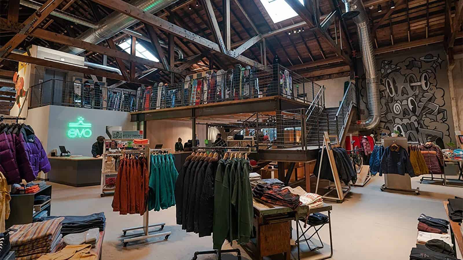 Clothing and outerwear displayed on racks inside a spacious, industrial-style Evo retail store—founded by skier Bryce Phillips—with exposed beams, mezzanine level, and a mural on the wall.