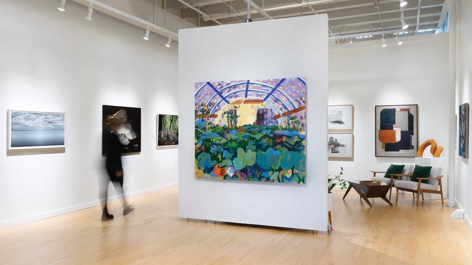 Art gallery interior with various paintings on white walls, wooden floor, a blurred person walking, mid-century modern chairs, and a small plant in the corner at J. Rinehart Gallery, a premier Seattle art gallery showcasing local art.