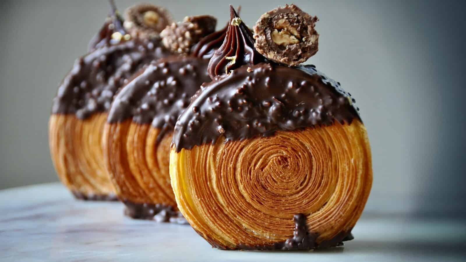 Three chocolate-dipped Petit Pierre pastry rolls with visible flaky layers, topped with a chocolate swirl and a round chocolate candy, arranged in a row on a light surface—truly the Best of the Best.