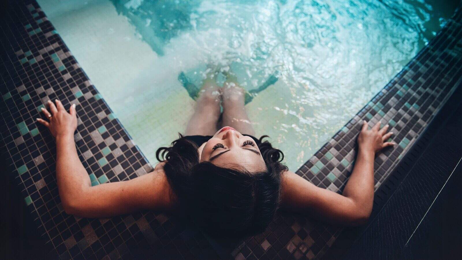 Unwind at Yuan Spa: a person relaxing in a tiled indoor pool, leaning back against the edge with arms outstretched and head tilted upwards, experiencing the best spa tranquility.
