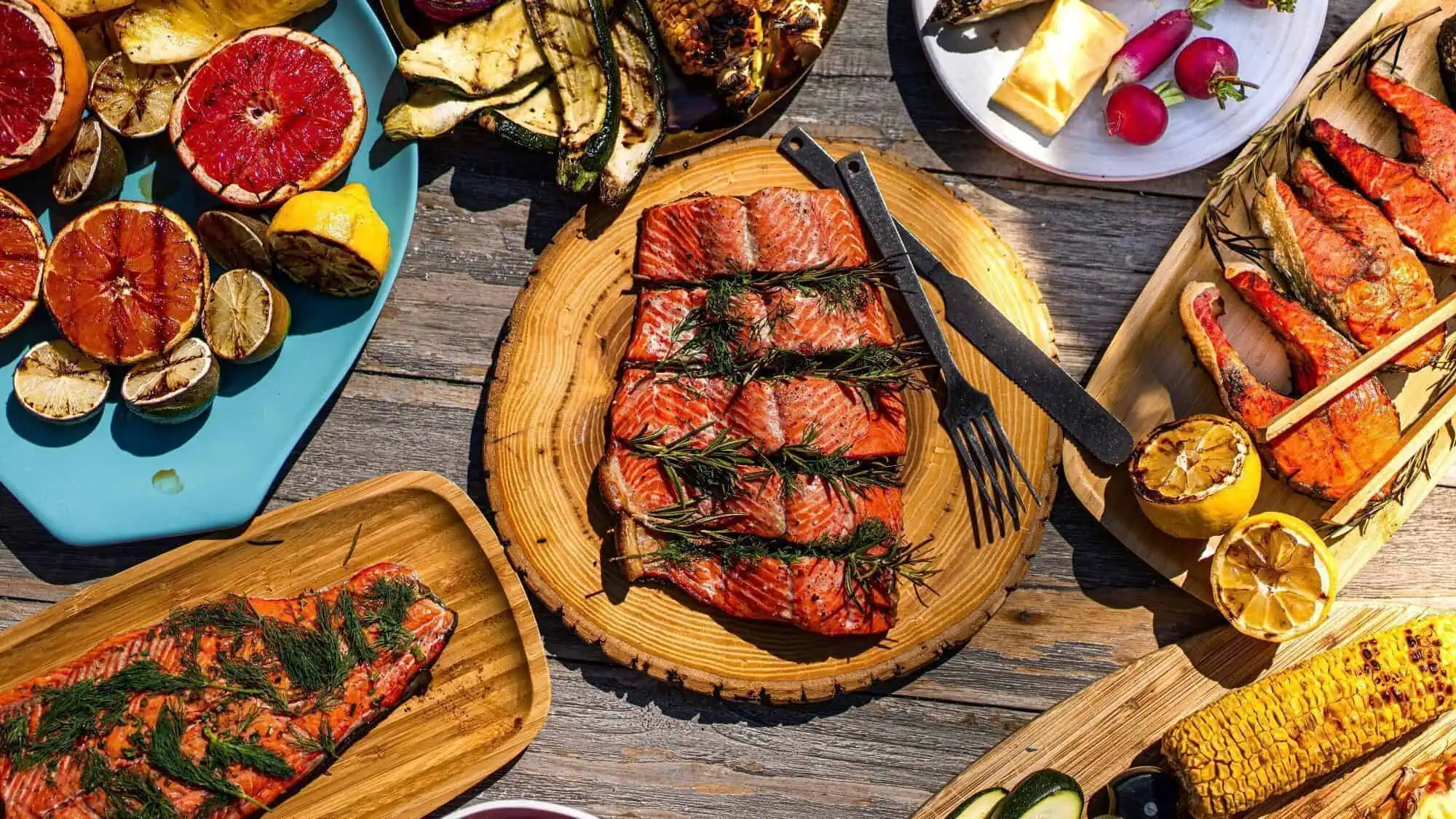Grilled salmon fillets with herbs on a wooden plate, surrounded by grilled vegetables, citrus, and corn on a rustic wooden table.