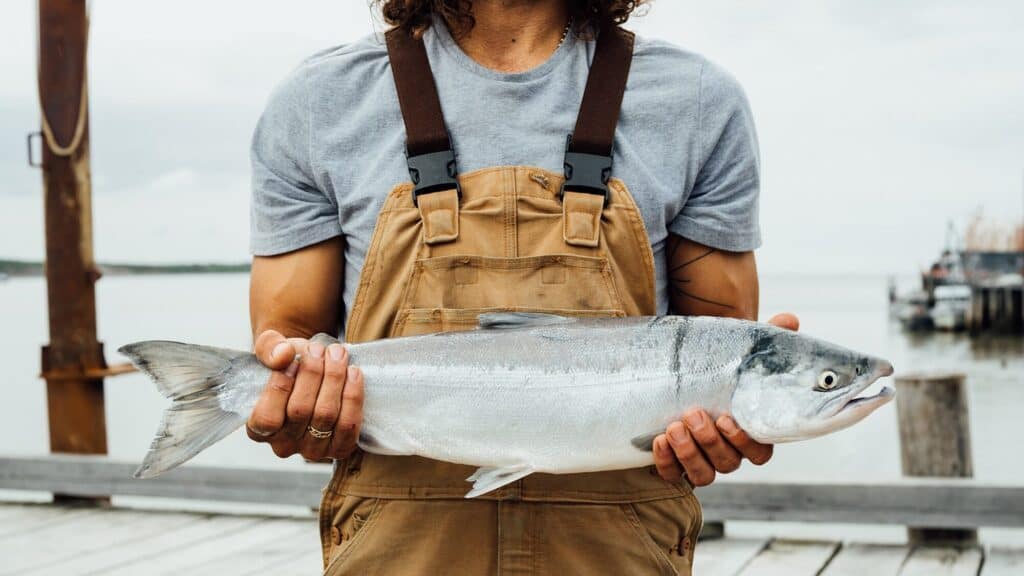 Person in brown overalls and gray shirt holding a large silver fish outdoors near water with a wooden dock in the background.