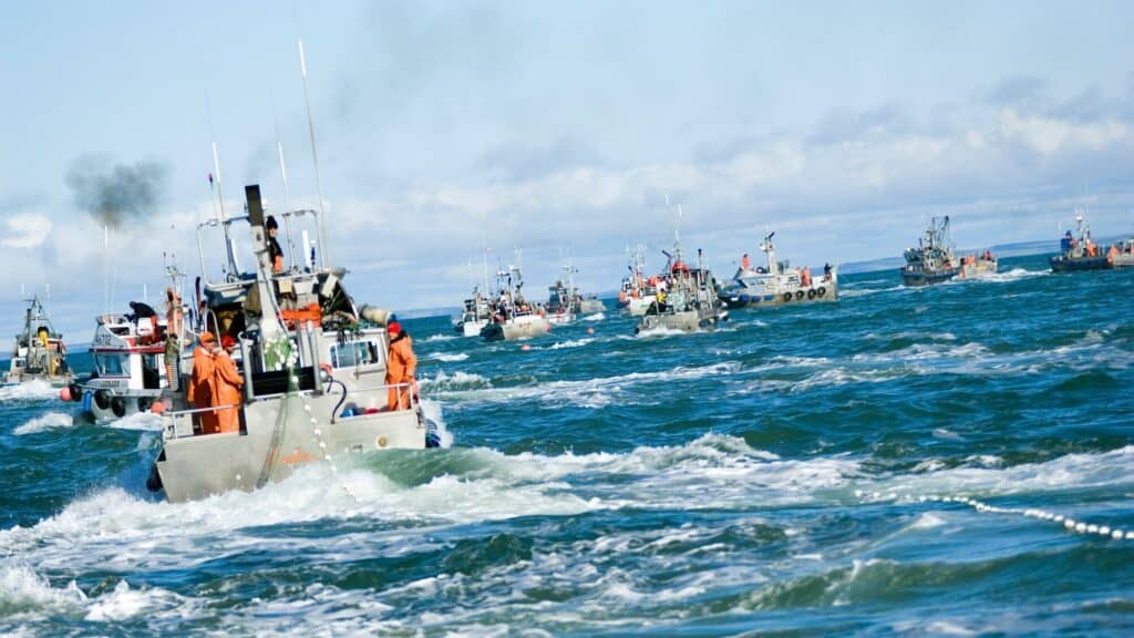 Several fishing boats with crew in orange gear navigate choppy blue waters under a partly cloudy sky, with more boats visible in the distance.