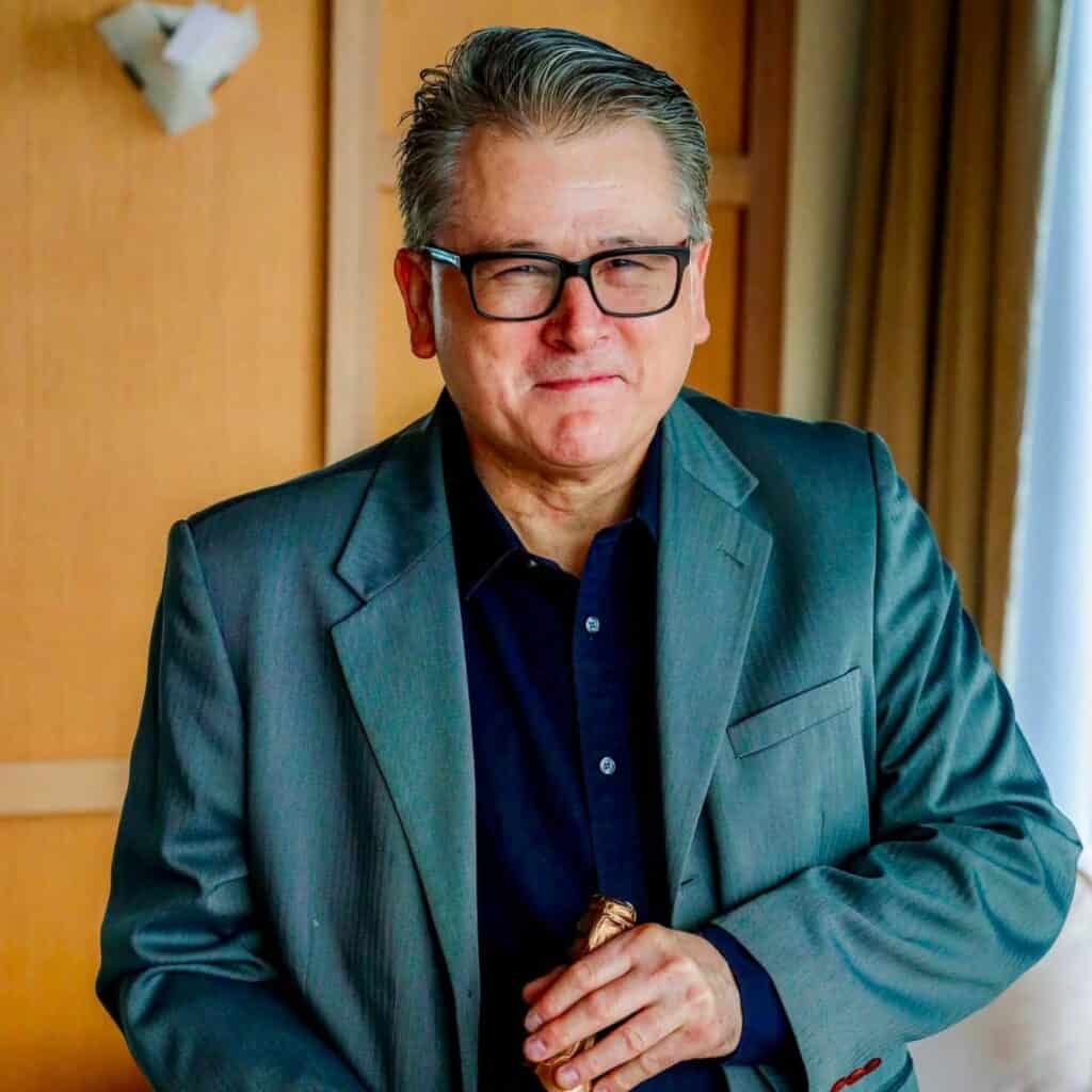 A middle-aged man wearing glasses and a grey suit stands indoors, holding a cigar and smiling slightly at the camera, ready to enjoy Thanksgiving dining with friends.