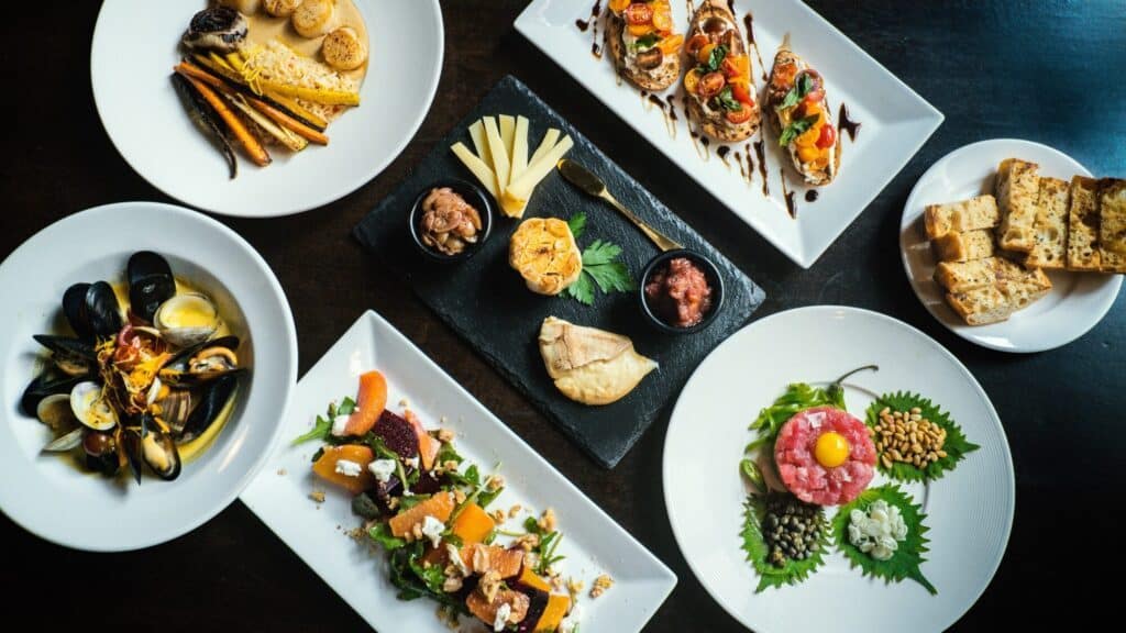 An overhead view of assorted gourmet dishes on white plates, including seafood, salads, bruschetta, cheese, bread, and garnished appetizers arranged neatly on a dark table.