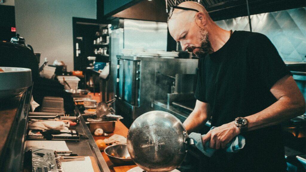 A man wearing a black t-shirt and sunglasses on his head washes a metal mixing bowl in a commercial kitchen.