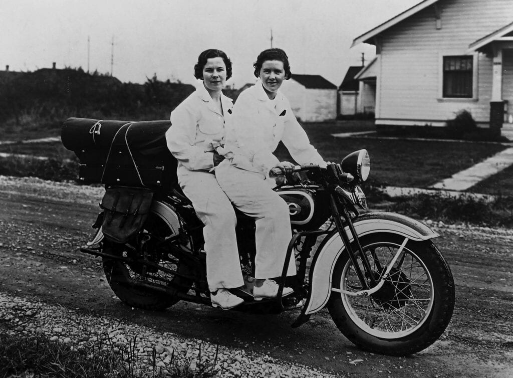 Two women in matching outfits sit on a motorcycle parked on a gravel road, capturing the spirit of Pacific Northwesterners, with houses and grassy yards visible in the background.