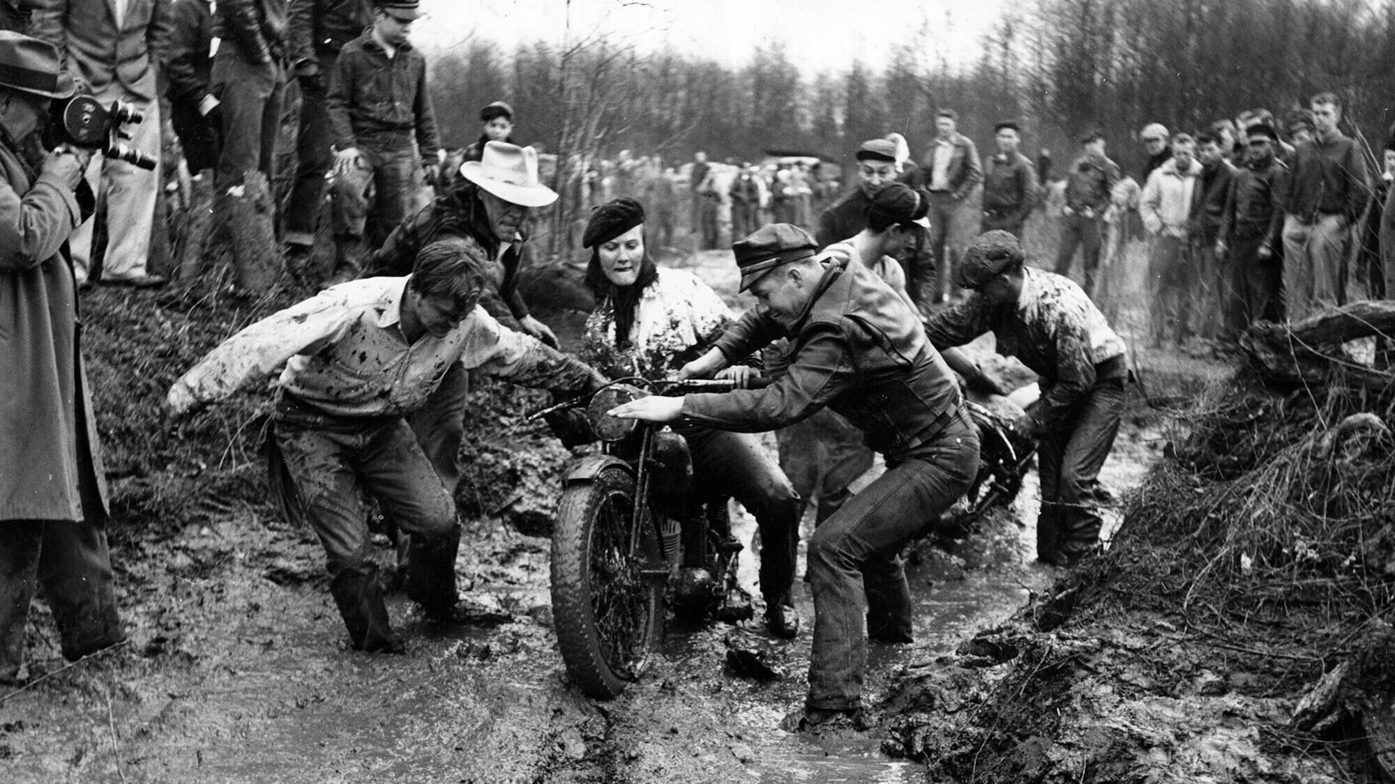 A group of Pacific Northwesterners helps push motorcycles through a muddy path while onlookers, some in raincoats and hats, observe in this classic outdoor scene, reminiscent of the adventurous spirit celebrated at MOHAI.