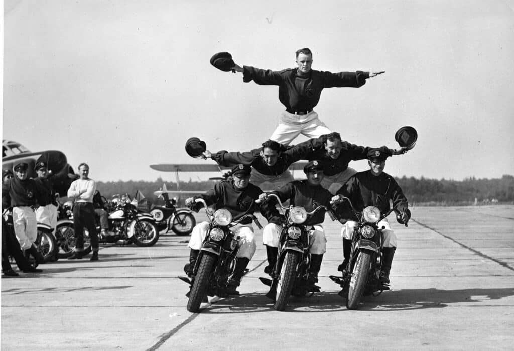 Five Pacific Northwesterners pose on four motorcycles at MOHAI, with three riding and two balancing on top, all holding hats, on an open airfield dotted with other motorcycles and a plane in the background.