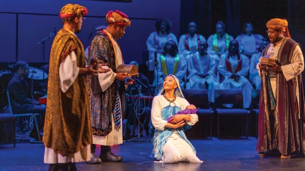 Four people in elaborate costumes perform a nativity scene on stage, one kneeling and holding a baby, while the choir looks on with joy—capturing the spirit of Merry Christmas Baby during memorable holiday events.