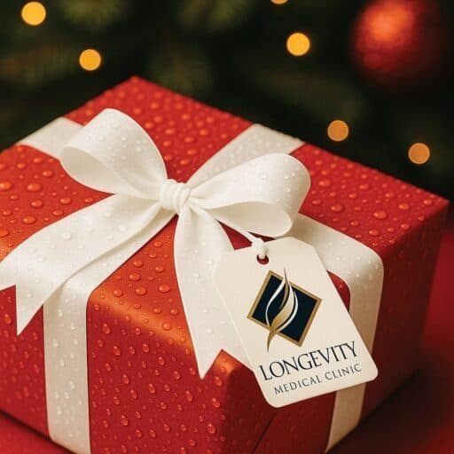 A red gift box with a white ribbon and bow sits on a surface. A tag displaying the Longevity Medical Clinic logo adds a thoughtful touch—perfect for holiday gift ideas. Blurred holiday lights glow in the background.
