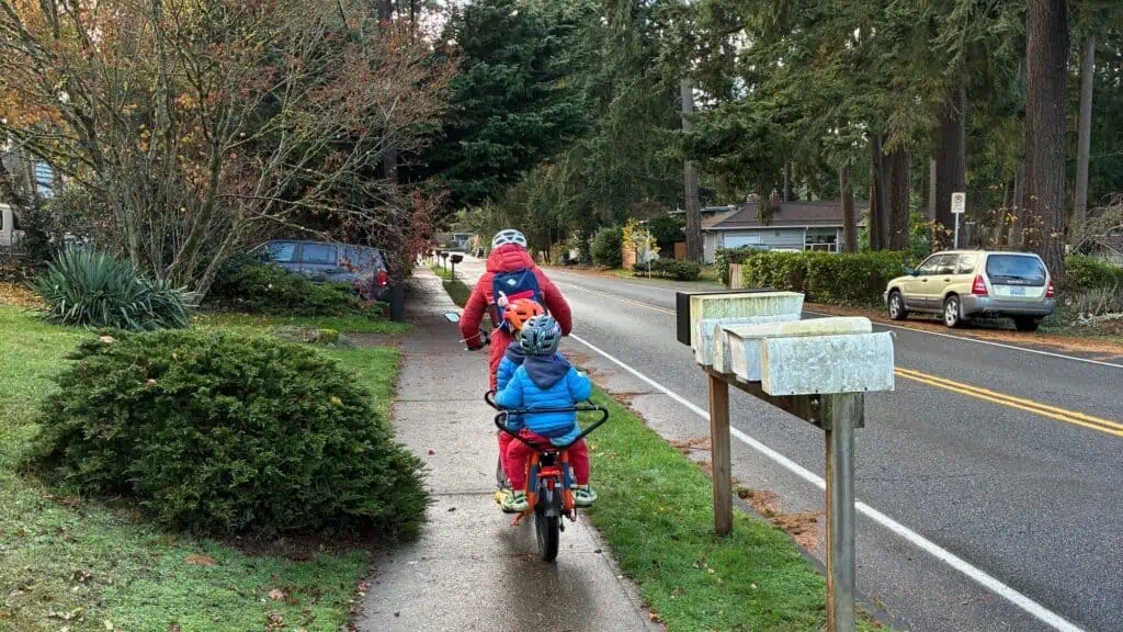 An adult and child wearing helmets and red jackets ride bicycles on a sidewalk next to a suburban street with mailboxes and parked cars.