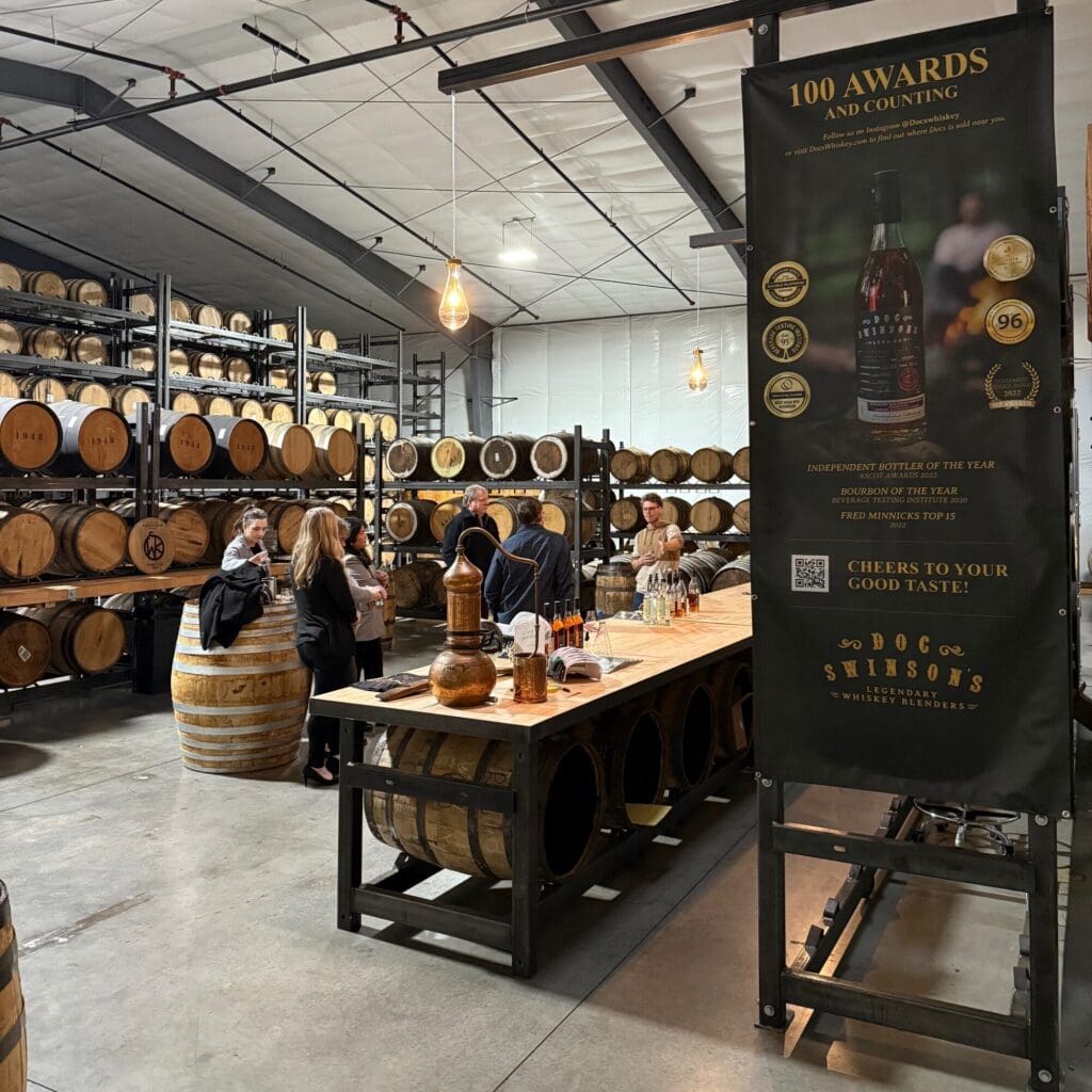 Lidé stojí kolem degustačního stolu ve skladišti lihovaru plném naskládaných dřevěných sudů; transparent zvýrazňuje ocenění a vyznamenání lihovaru.