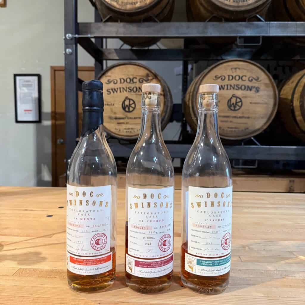 Tři částečně naplněné lahve Doc Swinson's Exploratory Cask Whisky Brand jsou vystaveny na dřevěném stole se sudy naskládanými v pozadí, které zachycují ducha objevování Little Whisky.